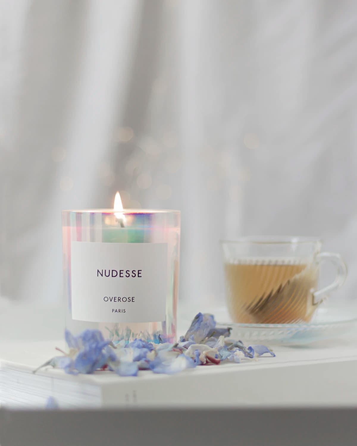 A lit candle beside a glass cup and blue flower petals.