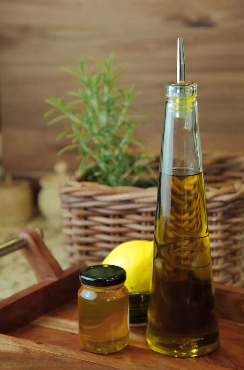A bottle of olive oil beside honey, lemon and rosemary on a wooden tray.