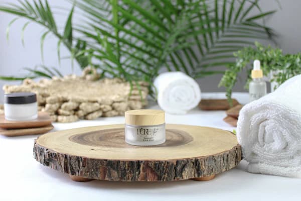 A small skincare jar on a wood slice beside rolled towels and greenery.